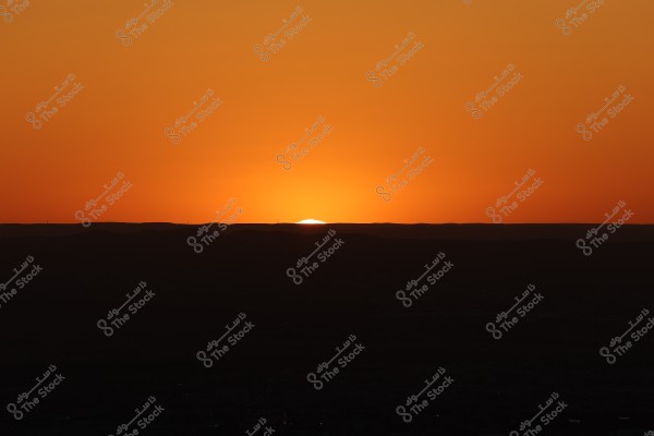 A sunset view with the sun setting behind the horizon, creating an orange sky. The land\'s silhouette forms a dark background at the bottom as the last bit of the sun disappears behind the line separating the sky and the earth.