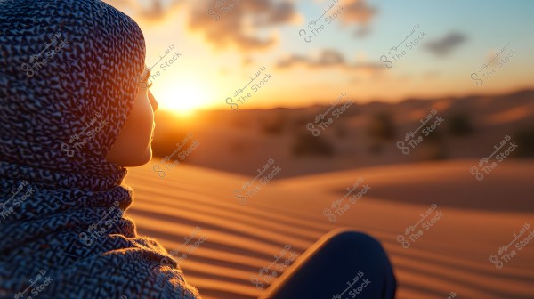 صورة لشخص يرتدي وشاحاً مزخرفاً يجلس في صحراء عند الغروب, حيث تملأ السماء السحب القليلة وتغمر الرمال الظلال البرتقالية الدافئة.