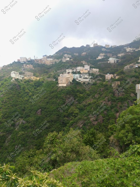 A landscape featuring a collection of houses scattered across lush green hills in a mountainous area. The sky is covered with light clouds. The buildings vary in color and are dispersed along the mountainside, indicating infrastructure adapted to the rugged terrain.