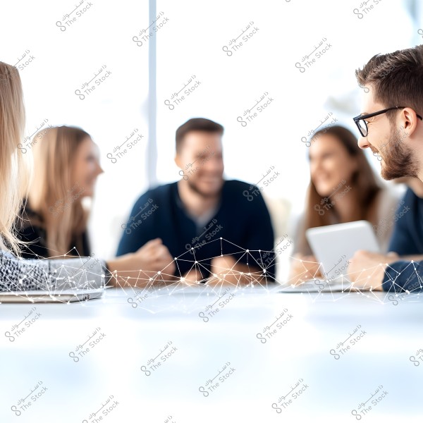 A group of people sitting together around a table in a brightly lit office environment. The image shows four people smiling and talking, with one holding a tablet. There is a floating digital network design on the table, adding a modern and technological feel.