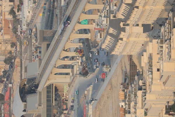 This is an aerial image of a modern city featuring a bridge with a train traveling on it, with distinctive architectural columns. Below the bridge, a road with cars moving in both directions is visible, along with residential and commercial buildings on the side.