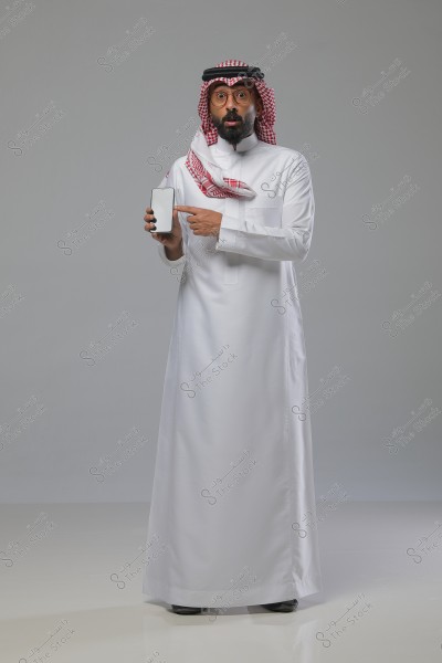 رجل يرتدي الثوب السعودي التقليدي والغترة الحمراء والبيضاء, يقف في وضعية بورتريه. يظهر الرجل وهو يحمل هاتفًا ذكيًا بين يديه ويشير إليه. خلفية الصورة باللون الرمادي.