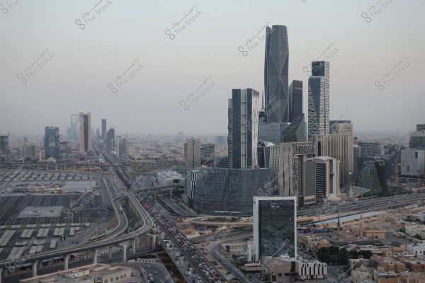 Panoramic view of the urban landscape in Riyadh, Saudi Arabia. The image features modern skyscrapers with distinctive architectural designs, including the prominent Kingdom Tower in the background. The scene includes winding highways filled with cars through the city, surrounded by rows of modern residential and commercial buildings.