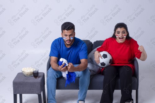 Portrait of a Saudi young man and woman watching a match and cheering their favorite teams cheerfully while having a snack on a blue background, the World Cup.