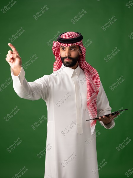 A portrait of a man wearing a white thobe and a red checkered headscarf (ghutra) with a black agal, standing in front of a green background. He appears to be pointing forward with his right hand while holding a tablet in the other hand. This attire is traditional in Saudi Arabia.