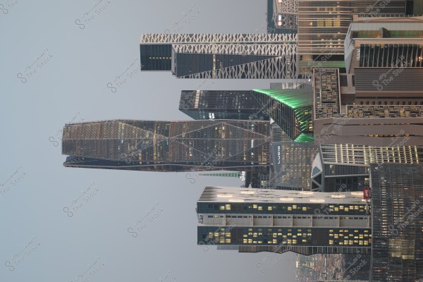 An urban scene depicting a collection of modern skyscrapers in Riyadh, Saudi Arabia. The buildings showcase intricate architectural designs, with some illuminated in green and blue lighting. The skyline appears in the background with a subdued color, as city lights cast a glow on the glass windows of the towers.
