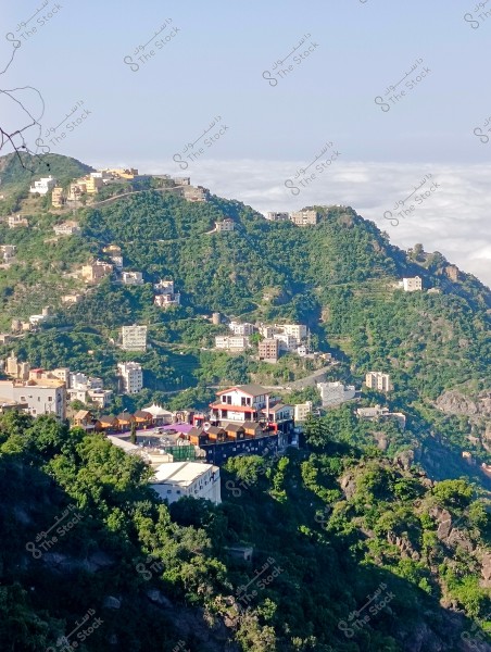 A stunning mountain view showcasing a city built on lush green slopes. The buildings are scattered haphazardly across the hillsides, with clusters of multi-story structures. In the background, clouds can be seen brushing against the mountain peaks, giving the impression that the city is perched above the clouds. Green vegetation covers most of the area, highlighting the beauty of the natural landscape.