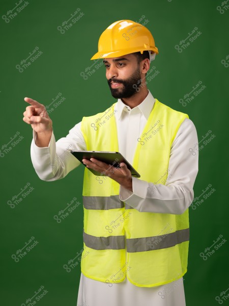 Image of a man wearing a yellow hard hat and a reflective safety vest over a white thobe, pointing with his hand while holding a tablet. The background is green, suggesting he is working in construction or engineering.