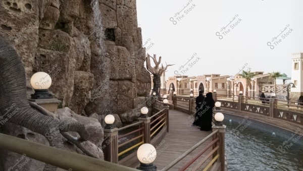 A scene of a water pathway beside a large rock formation in a setting resembling a traditional village. Glowing lanterns line the wooden walkway over the water. The image shows two trees with bare branches and several people in black cloaks walking along the path.
