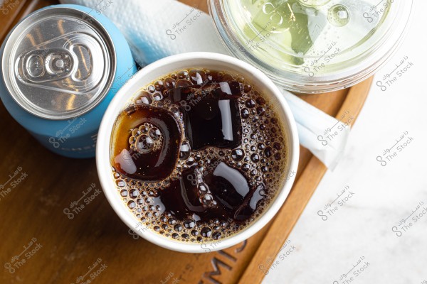 كوب من القهوة المثلجة مع قطع ثلج بجانب عبوة زرقاء وزجاجة بها غطاء شفاف على صينية خشبية.