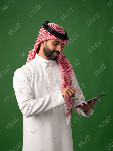 صورة لرجل يرتدي الثوب السعودي التقليدي والغترة والعقال. يقف أمام خلفية خضراء بينما يستخدم جهاز لوحي. يرتدي الرجل ثوباً أبيض ويبدو مركزاً على الجهاز.