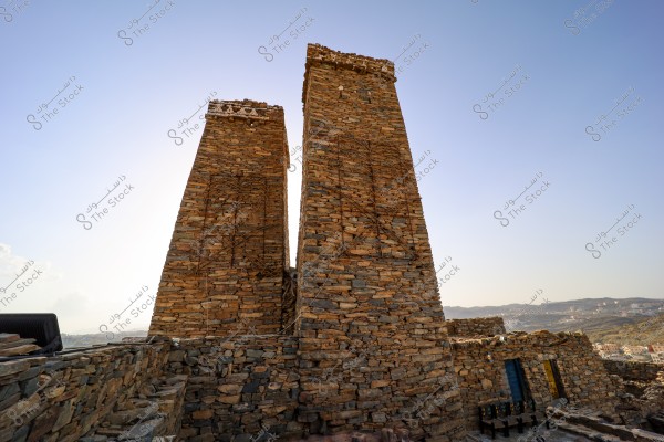 The image shows traditional stone towers made of rock in a mountainous area under a clear blue sky. The architectural design reflects an ancient style, and in the background, there is a scenic view of mountains and some buildings.