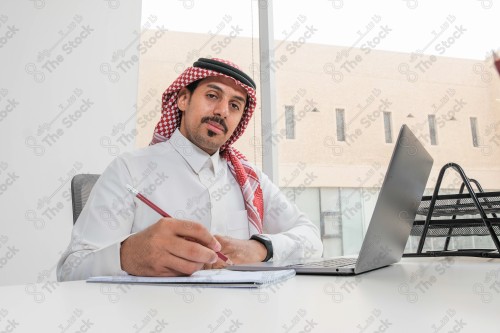 رجل سعودي يرتدي الزي السعودي التقليدي يعمل في مكتب بواجهة زجاجية ويستخدم قلم رصاص لتدوين الملاحظات