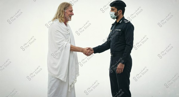 Two men shaking hands, one in a white Ihram garment with long blond hair, and the other in a dark police uniform with a face mask. The background is plain white.