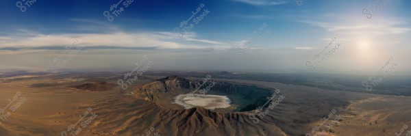 Aerial view of the \"Al Wahbah\" volcanic crater in Saudi Arabia, featuring a white crater floor amidst a desert surrounded by rocky terrain. The sky is blue with some scattered clouds, and the sun shines from behind the horizon, casting a warm light on the scene.