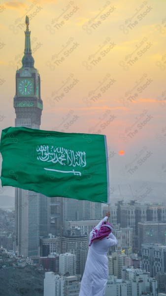 A snapshot of a Saudi man wearing the traditional Saudi dress, carrying the Saudi flag over a mountain in the Makkah Al-Mukarramah region, and the sky appears clear, the royal clock tower building in the Grand Mosque, buildings and landmarks, the Grand Mosque of Mecca.