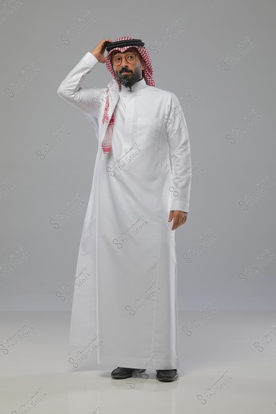 A portrait of a man wearing traditional Saudi attire, consisting of a white thobe and a red and white keffiyeh. The man stands in a formal pose, holding the edge of his keffiyeh, with a calm expression. The background is a simple gray.