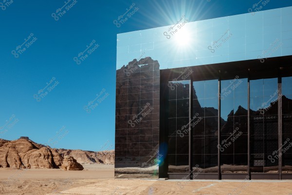 A building with a glass facade reflecting rock formations and a clear blue sky in a desert landscape. The sun shines at the top, adding a gleaming effect to the facade.