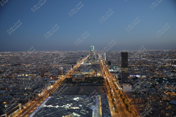 Aerial view of Riyadh\'s skyline at night, showcasing a long illuminated road running through the city leading to the Kingdom Tower, distinguished by its unique design and green lighting. Surrounding are buildings and lit streets, reflecting the urban landscape of the city.