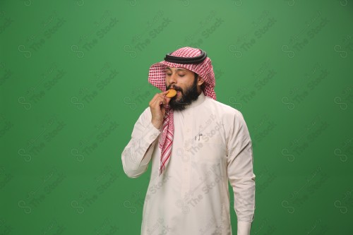 شاب سعودي على خلفية خضراء يرتدي الثوب و الشماغ، يقوم بعمل حركات بيده،يمسك الكعك و يأكله ، احتفالات العيد