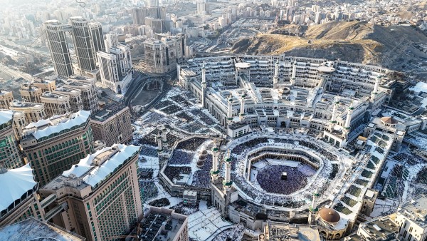 An aerial view of the area surrounding the Masjid al-Haram in Mecca, Saudi Arabia. The image prominently displays the Grand Mosque, including the Kaaba surrounded by a throng of worshippers and pilgrims. The scene is encircled by many tall buildings and hotels showcasing modern architectural designs, with the mountainous landscape of the city visible in the background under bright daylight.