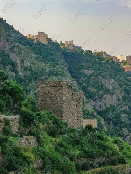 A natural landscape of a mountain covered with dense green vegetation, featuring a traditional stone structure in the center. In the background, several buildings are visible on the peaks of the surrounding mountains. The sky is clear blue with a few light clouds.
