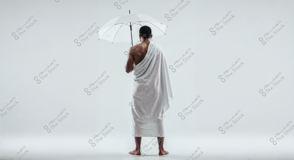 An image of a man standing with his back to the camera, wearing a white wrap similar to Ihram clothing, holding a white umbrella. He appears to be in a minimalist setting with a white background.