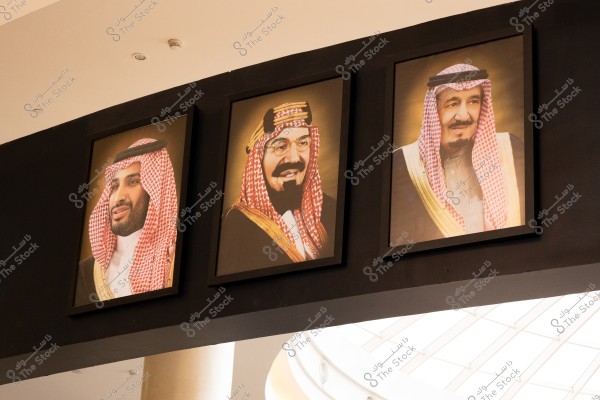 ثلاث صور شخصية مؤطرة معروضة على جدار بلون داكن. الصور تظهر ثلاثة رجال يرتدون الزي التقليدي السعودي, وكل صورة تحتوي على رجل يرتدي ثوباً أبيض مع شال أحمر وأبيض. الخلفية لأصفر فاتح وتحتوي الصور على أضواء من المبنى في الأعلى.