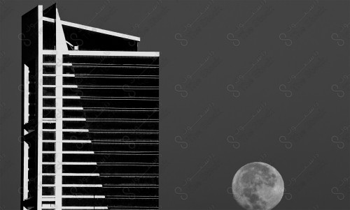 A black and white shot of one of the buildings in Riyadh at night, and the moon appears full behind it.