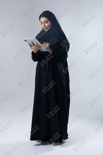 A portrait of a woman wearing a black abaya and hijab, standing while attentively looking at a tablet. She holds the device in her right hand and uses a digital pen with her left hand. The background is gray and empty, making the figure stand out distinctly. The image conveys a sense of technology and modernity.