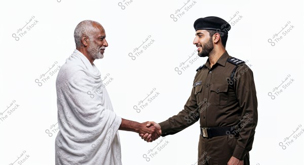 An image of two men shaking hands. The first man is elderly and wearing a white ihram, suggesting he may be on a pilgrimage for Hajj or Umrah. The second man is wearing a formal brown uniform, indicating he might be part of the military or police, possibly of Saudi origin due to the style of the uniform.