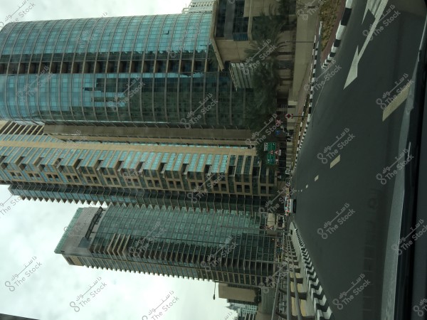 Image showing tall skyscrapers in a modern city, featuring contemporary architectural design with glass facades. In the foreground, a wide road with clear road markings and palm trees on the right side. The sky is overcast, giving the scene a slightly gray tone.