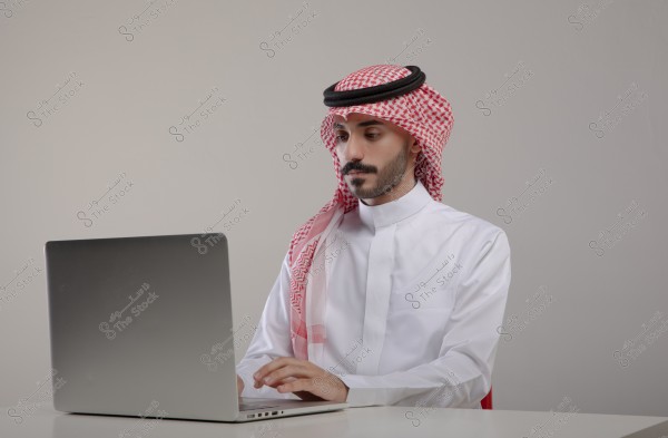 An image of a man wearing a white thobe, a red shemagh, and a black agal, sitting in front of a silver laptop. He appears to be working or focused on the screen. The background is neutral and simple.