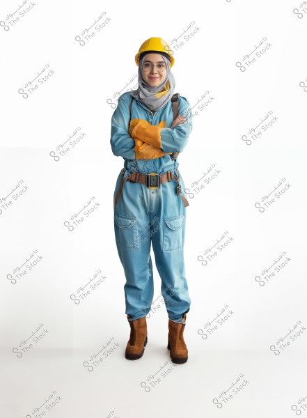 The image depicts a woman dressed in a blue work uniform that covers her entire body. She wears glasses, a yellow hard hat, and orange gloves. The woman stands with her arms crossed and is wearing brown boots. The background is white, emphasizing the details of the attire, which may be related to a technical or industrial profession.