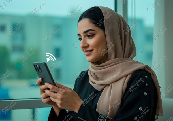 صورة لامرأة ترتدي حجابًا بلون بيج وعباءة سوداء, تجلس بجوار نافذة زجاجية ممسكة بهاتف ذكي. يبدو المبنى غير واضح في الخلفية. تعابير وجهها توحي بالسعادة.