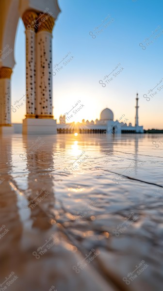 Image of a prominent mosque at sunset, showcasing its white structure with traditional Islamic design, a large dome, and a tall minaret. The golden light reflects on the wet ground surface, adding an aesthetic touch to the scene. Decorative architectural details with gold embellishments are visible on columns in the foreground.