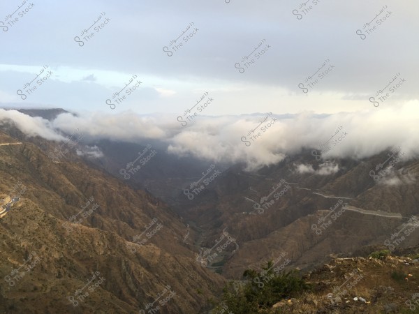 An image showcasing a natural landscape of a high mountain with a winding road running through it. White clouds cover parts of the mountain, and beneath them, deep valleys are visible. Greenery is scattered on the rocky hills, with a light blue sky above.