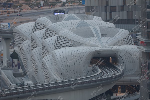 A modern train station with a complex architectural structure, showing train tracks entering a tunnel beneath a wavy roof. The building\'s exterior is adorned with mesh patterns and decorated with white metal panels, extending alongside a highway visible in the background with some cars and urban infrastructure.