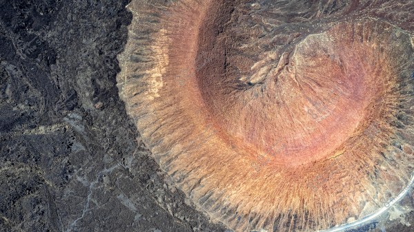 An aerial view of a volcanic crater in a desert landscape. The crater is centrally located in the image and is characterized by a reddish-brown color that stands out prominently against the surrounding dark rocks. The irregular edges and terrain details of the crater are clearly visible, conveying a sense of depth and unique geological formation.