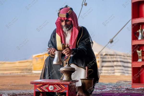 A picture of a man sitting on a carpet in a setting that resembles a traditional tent. The man is wearing traditional Saudi attire with a red keffiyeh and a black agal. In front of him is a decorated wooden table with traditional copper vessels, and behind him, there\'s a large tent. The scene reflects Bedouin culture.