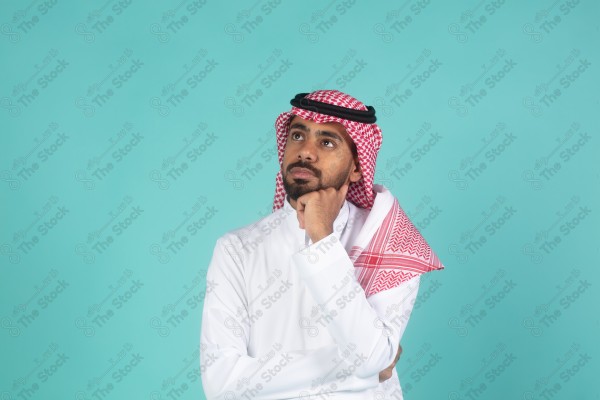 بورتريه رجل سعودي يرتدي الثوب يقف أمام خلفية تركوازية ، تعابير وجه تدل على التساؤل والحيرة والتفكير ، لحظة اتخاذ قرار ، صورة شخصية