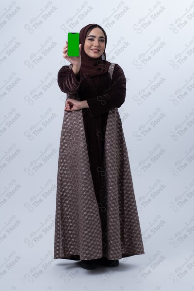 A young Saudi woman wearing an abaya and hijab, holding a green-screen phone