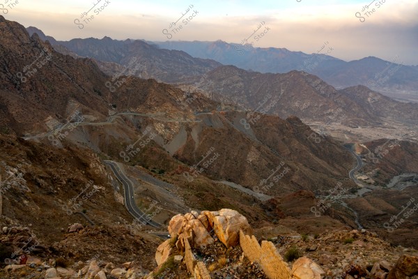 A natural landscape of towering mountains in the Asir region of Saudi Arabia, featuring winding roads ascending the mountains, with prominent rock formations in the foreground. The horizon is filled with overlapping mountain ranges beneath a cloudy sky.