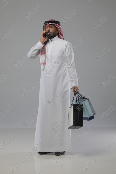 Image of a man wearing a Saudi thobe and a red ghutra, talking on a mobile phone and holding several shopping bags in different colors. He appears relaxed against a grey background.