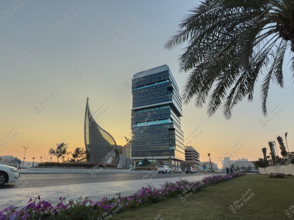 صورة لشارع في مدينة حديثة عند الغروب, يظهر فيها مبنى زجاجي طويل بتصميم معاصر ونصب فني على شكل قارب. هناك حركة بسيطة للسيارات على الطريق, وأشجار النخيل تزين المشهد مع أزهار على جانبي الطريق. السماء تتلون بألوان الغروب الدافئة.\r\n\r\n-