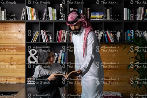 رجل سعودي مبتسم يرتدي الزي السعودي التقليدي يقف بجانب فتاة سعودية ويتصفحون أحد الكتب في المكتبة