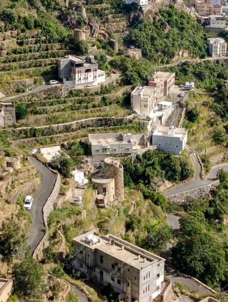Aerial view of a mountainous village featuring lush green agricultural terraces. The traditional houses are situated on the hillsides and slopes with varying designs. There are two narrow roads winding through the village, extending across the mountains. Some buildings are modern, and there is a traditional circular stone tower. The area is rich with vegetation and trees.