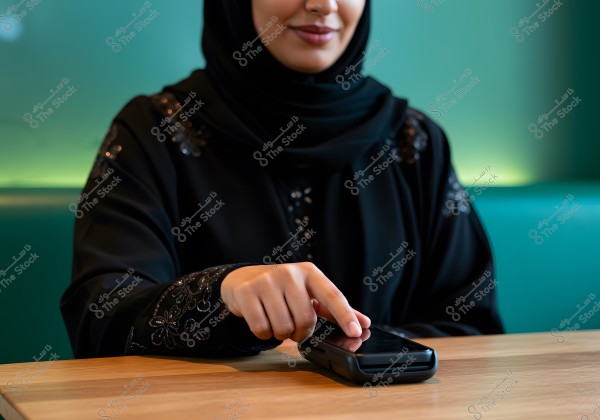 An image of a woman wearing a decorated black abaya and hijab, touching an electronic payment device with her hand in a well-lit indoor setting with a green background. The focus is on the upper half of the woman and the motion of her hand.