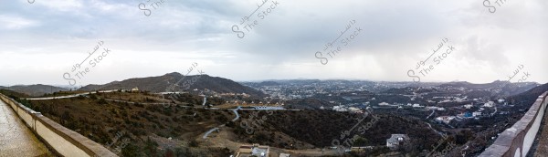Panoramic view of a mountainous area in Taif, Saudi Arabia. The image shows a winding road with a fence on the left, and scattered houses on the hills. The sky is cloudy, with mist covering parts of the mountains in the background.