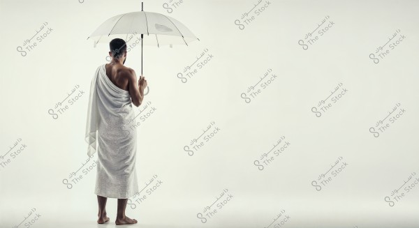 A man is standing in a portrait, wearing the white Ihram clothing, consisting of two unstitched pieces of white cloth wrapped around his body. He is holding a white umbrella in his right hand. The background is a plain white, emphasizing the Ihram attire and the umbrella. The image conveys a serene and elegant appearance.
